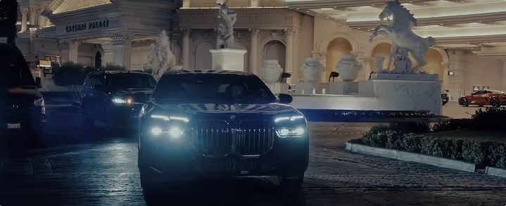Front view of a black luxury car with bright headlights driving at night near a building adorned with horse statues.