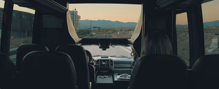 View from backseat looking forward inside a vehicle driving on a road with mountains visible through the windshield.