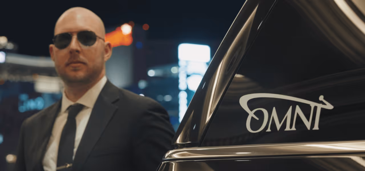 A private Omni chaffeur in a black suit and tie wearing sunglasses standing next to a black car with an OMNI logo on the window at night.